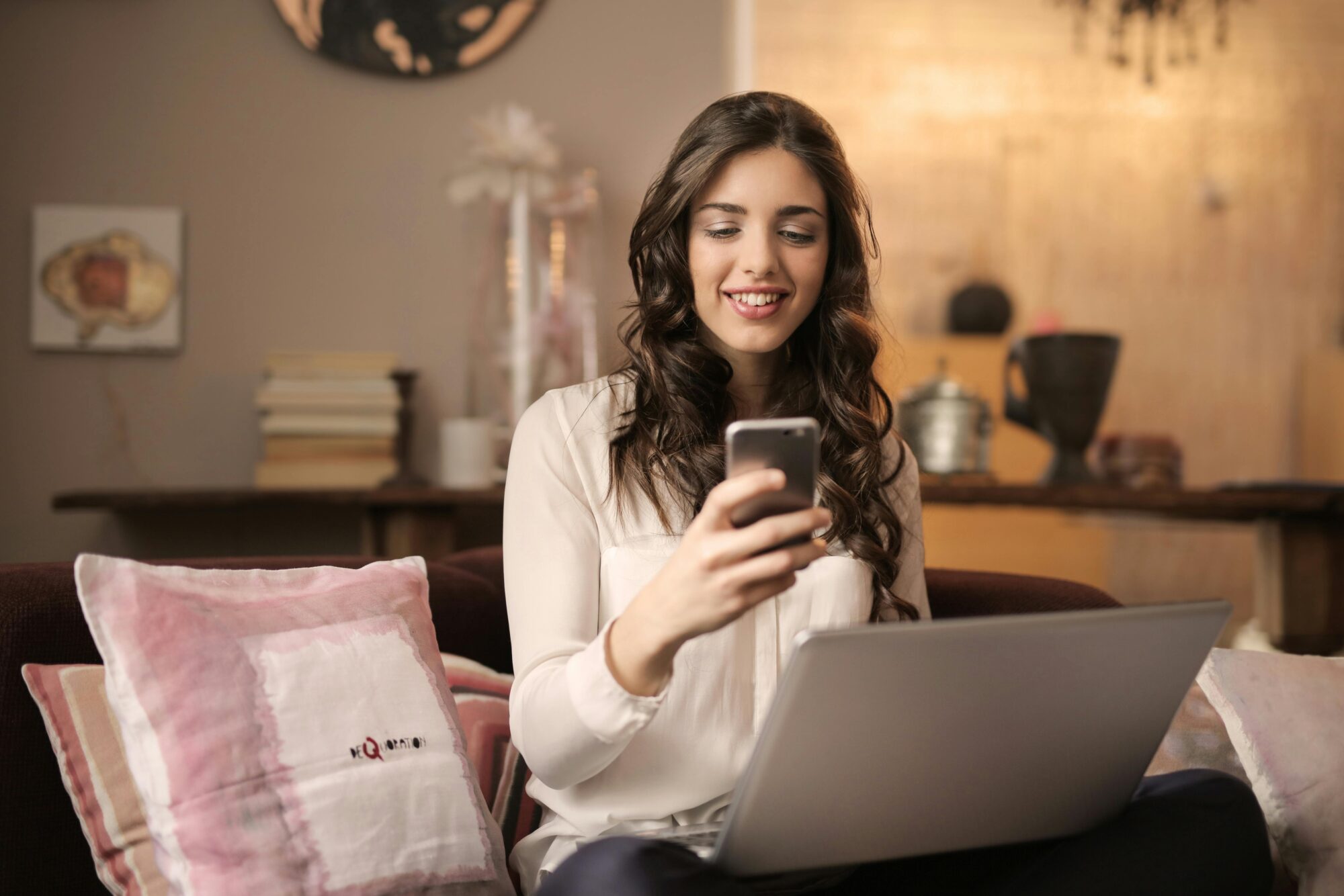 Woman sitting on sofa while looking at phone with laptop on lap