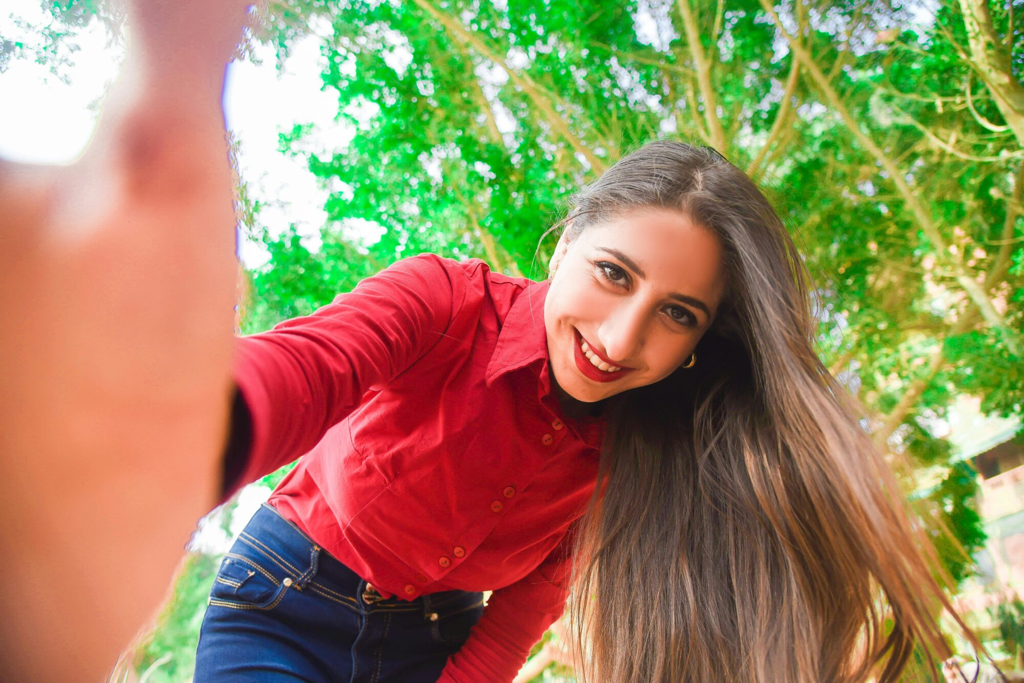 Smiling woman in red shirt and blue jeans taking selfie under green leaved tree